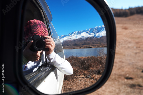 Reflection of a photographer woman with a camera in the rear view mirror of a car in nature mountain.