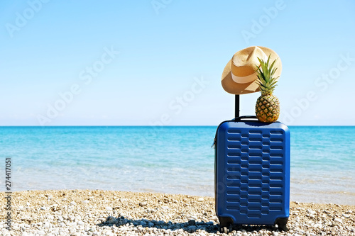 Blue hardshell carry-on roller luggage standing with straw hat & pineapple on sandy beach. Hard plastic suitcase on wheels w/ extended handle. Summer vacation concept. Copy space background, close up.