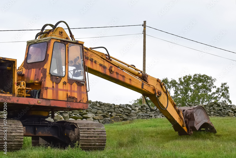 Big yellow excavator on a green hill near a stone fence. Concept: Road and construction work. Big yellow excavator on a green hill near a stone fence. Concept: Road and construction work.