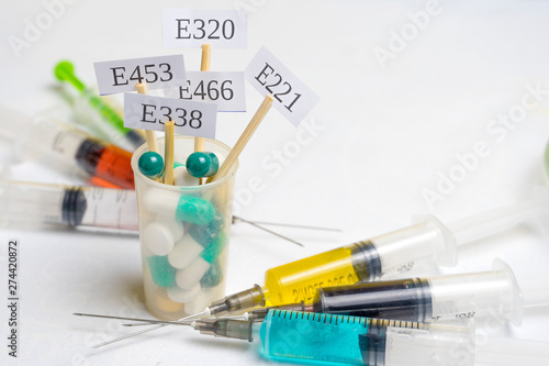 Harmful food additives. In a glass with multi-colored pills are plates with the code E-supplements. Nearby are syringes with nitrates, chemicals and additives