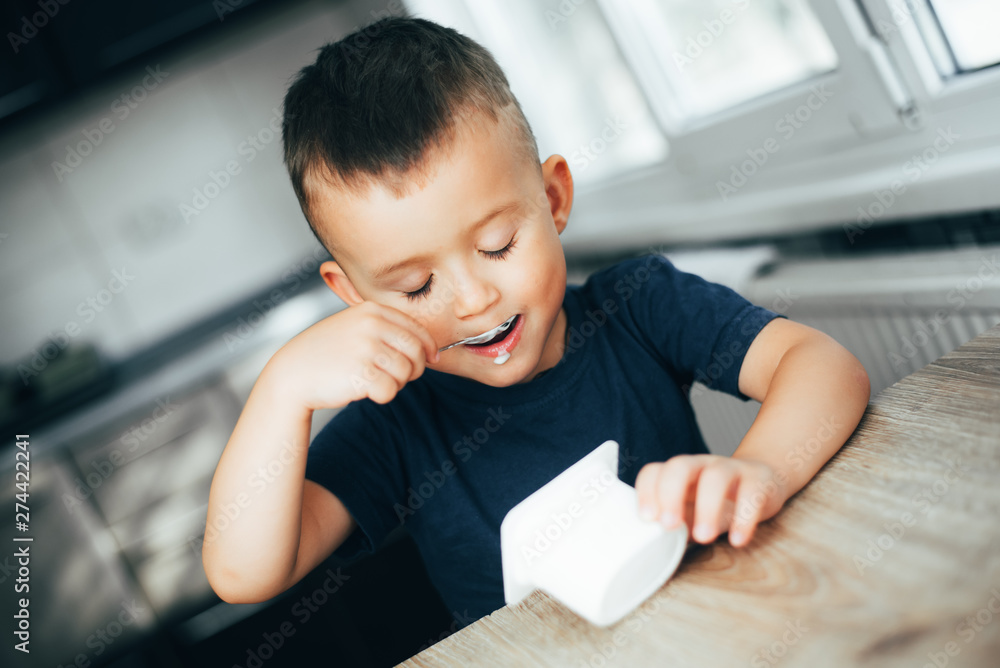 Cute boy in the kitchen eating yogurt from a white container for yogurt, there is a place for advertising Cute boy in the kitchen eating yogurt from a white container for yogurt, there is a place for advertising