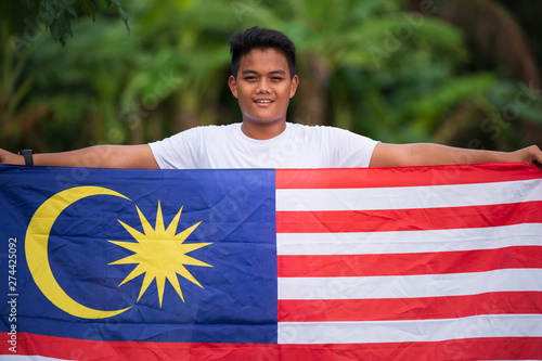 Portrait of young Malay Asian man standing, holding and waving Malaysian flag or known as Jalur Gemilang, happy, celebrating Malaysia National Independence's Day, copy space.