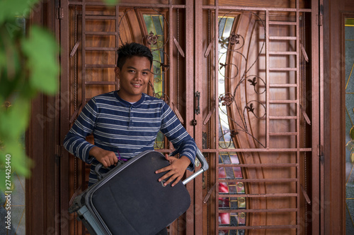 Portrait of young Asian man standing and holding luggage bag with house door background, leaving for holidays, traveling concept, outstation work, long trip.