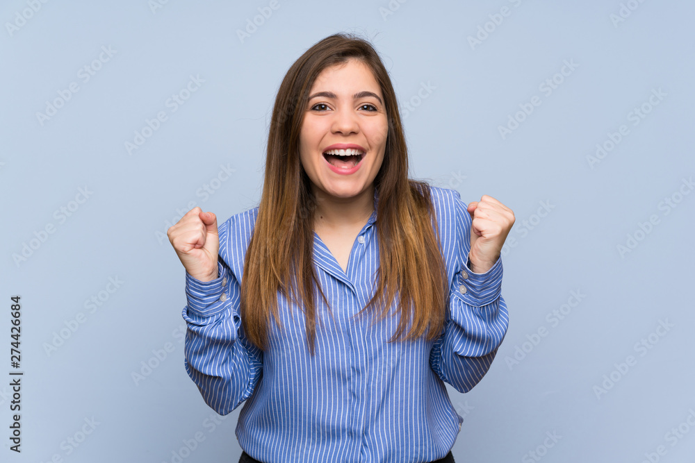 Young girl over isolated blue wall celebrating a victory in winner position Young girl over isolated blue wall celebrating a victory in winner position