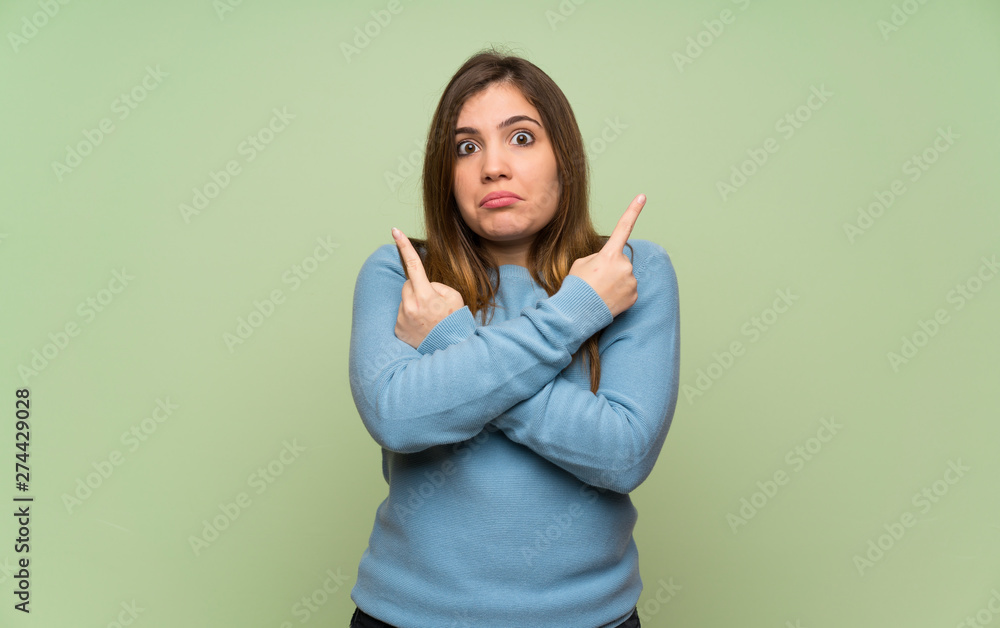 Young girl over green wall pointing to the laterals having doubts Young girl over green wall pointing to the laterals having doubts
