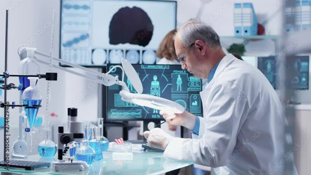 Mature researcher or doctor using a big industrial magnifying glass in modern secret laboratory Mature researcher or doctor using a big industrial magnifying glass in modern secret laboratory