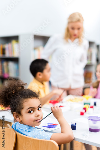 selective focus of cute black girl looking at camera near kids and woman