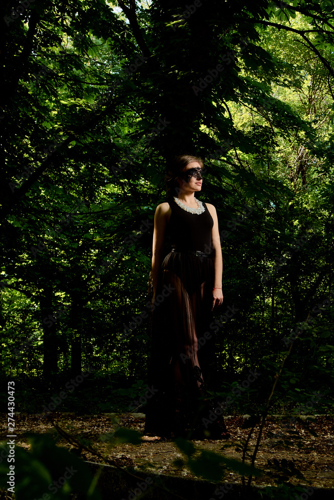 Young attractive woman in the sexy see-through, transparent, transpicuous, diaphanous, clear black dress and black lace masquerade venetian face mask posing outdoor at the green alley in the old park. Young attractive woman in the sexy see-through, transparent, transpicuous, diaphanous, clear black dress and black lace masquerade venetian face mask posing outdoor at the green alley in the old park.