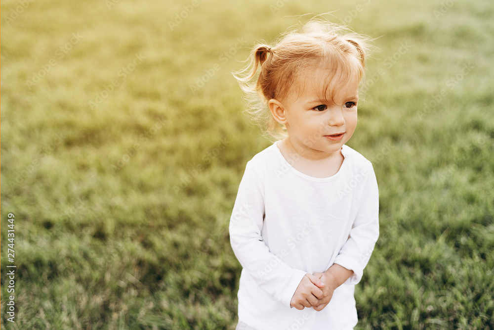 little baby girl with blond hair and two tails, wearing a white T-shirt standing on green grass in nature, smiling little baby girl with blond hair and two tails, wearing a white T-shirt standing on green grass in nature, smiling