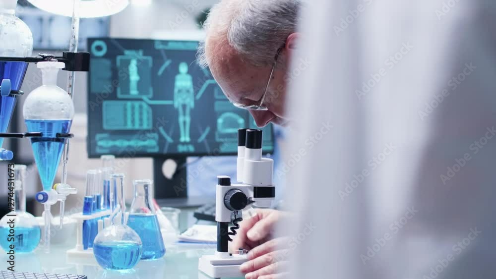 Close up dolly shot of mature chemist researching using a microscope. In the background - secret high end modern facility Close up dolly shot of mature chemist researching using a microscope. In the background - secret high end modern facility