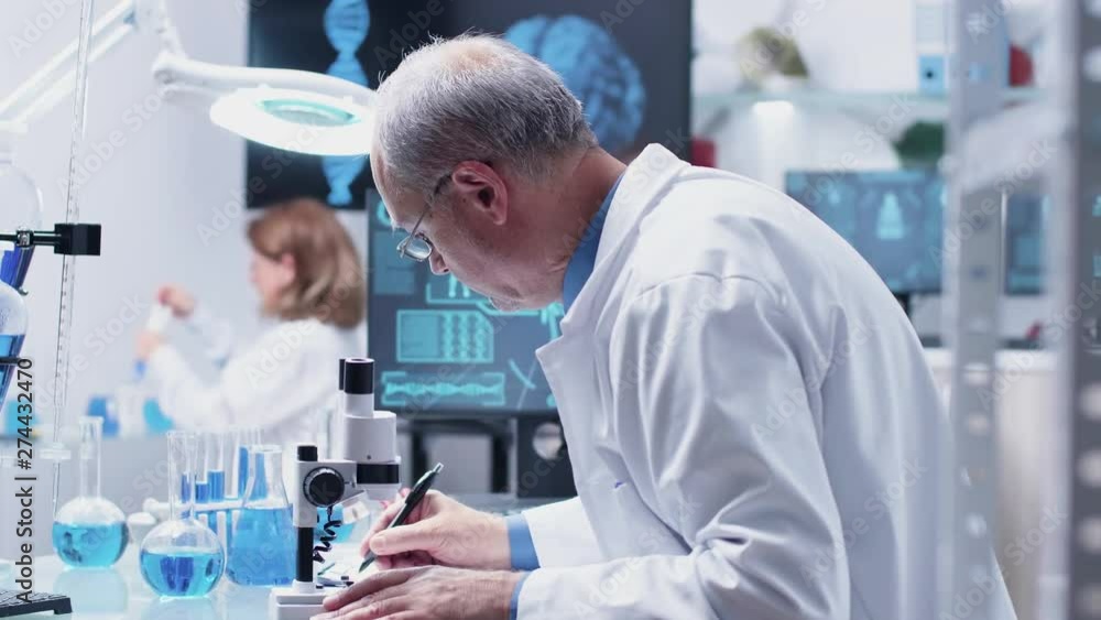 Dolly close up shot of mature chemist using a microscope and taking notes in a clipboard Dolly close up shot of mature chemist using a microscope and taking notes in a clipboard