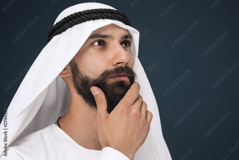 Half-length portrait of arabian saudi businessman on dark blue studio background. Young male model standing and looks thoughtful. Concept of business, finance, facial expression, human emotions. Half-length portrait of arabian saudi businessman on dark blue studio background. Young male model standing and looks thoughtful. Concept of business, finance, facial expression, human emotions.