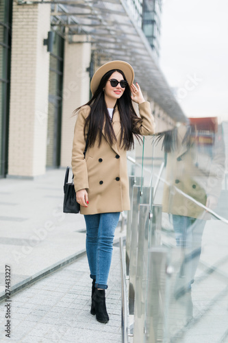 Young cheerful woman in hat and coat standing on balcony and smiling outdoors. Beautiful and fashionable hipster girl in eyeglasses touching hat and posing on city street. Woman in modern city outfit