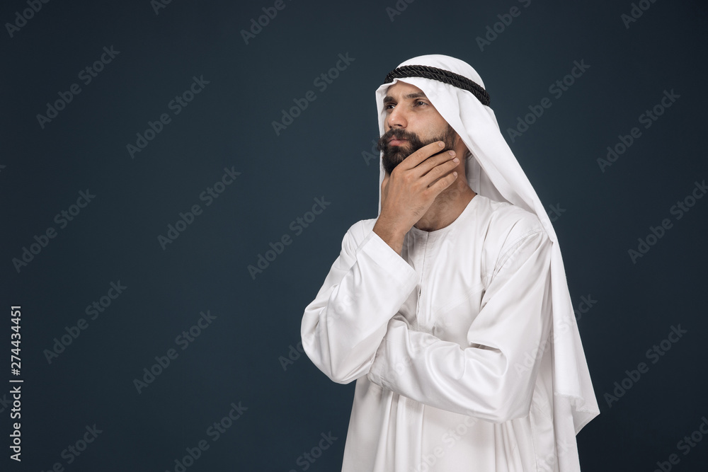 Half-length portrait of arabian saudi businessman on dark blue studio background. Young male model standing and looks thoughtful. Concept of business, finance, facial expression, human emotions. Half-length portrait of arabian saudi businessman on dark blue studio background. Young male model standing and looks thoughtful. Concept of business, finance, facial expression, human emotions.