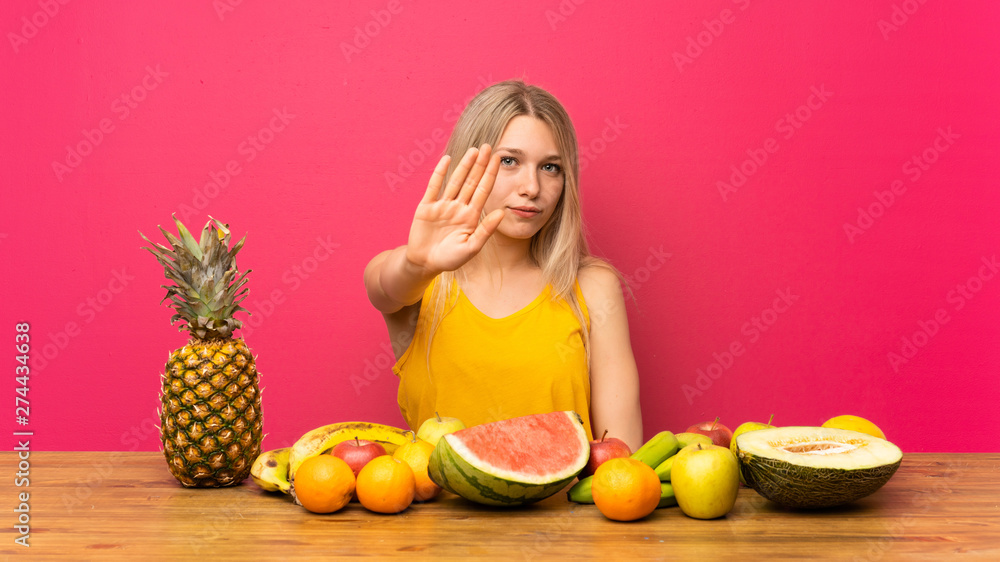 Young blonde woman with lots of fruits making stop gesture with her hand Young blonde woman with lots of fruits making stop gesture with her hand