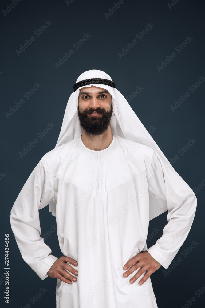 Half-length portrait of arabian saudi businessman on dark blue studio background. Young male model standing and smiling. Concept of business, finance, facial expression, human emotions. Half-length portrait of arabian saudi businessman on dark blue studio background. Young male model standing and smiling. Concept of business, finance, facial expression, human emotions.