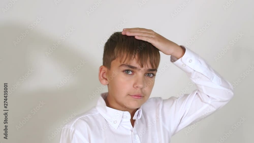 A ten-year-old boy in a white shirt stands against a white wall, he looks at the camera. Slow motion.