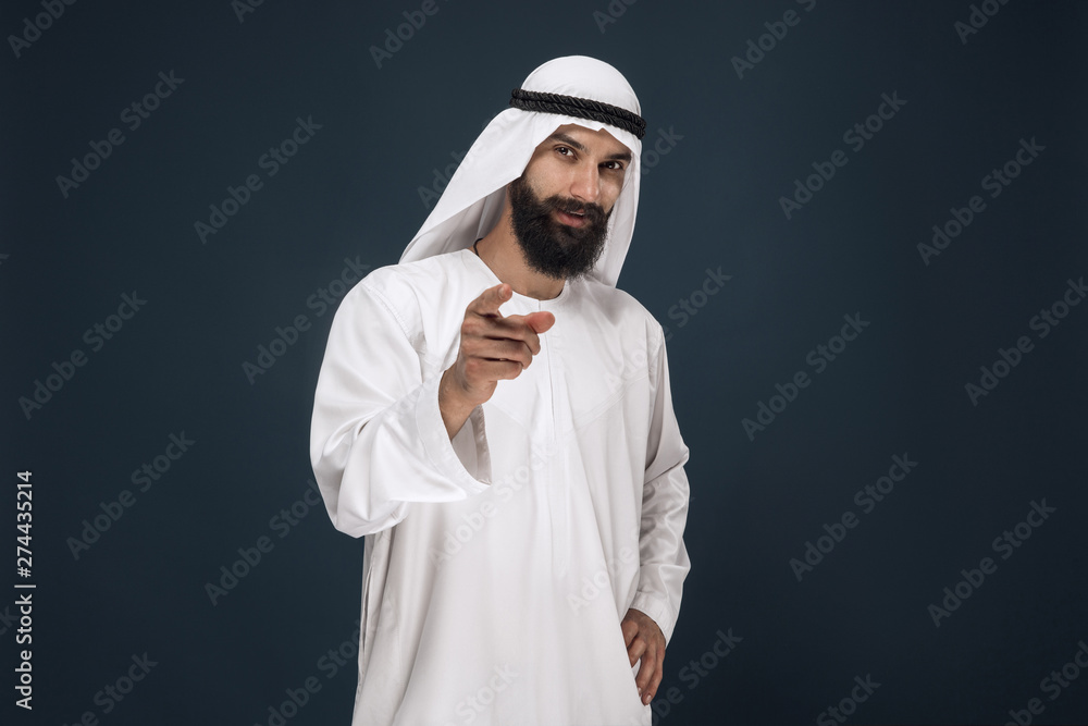Half-length portrait of arabian saudi businessman on dark blue studio background. Young male model smiling and pointing or choosing. Concept of business, finance, facial expression, human emotions.