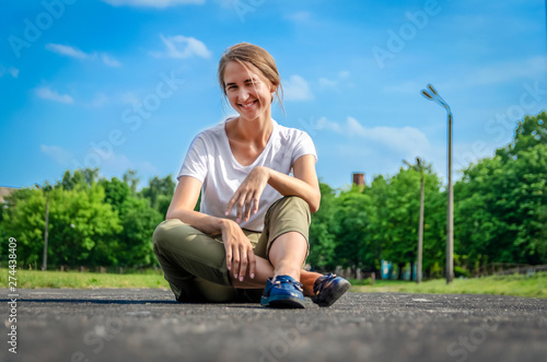 Beautiful brown-haired girl in a white T-shirt and khaki pants is sitting on the cement path of the stadium against the blue sky on a sunny evening
