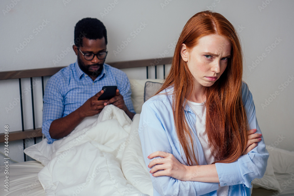 Fototapeta premium redhaired ginger caucasian happy female and multi-ethnic afro man together lying in bed bedroom looking at smartphones texting.Family conflict