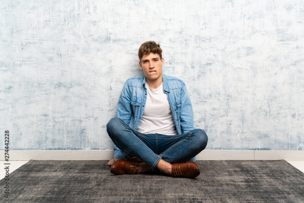 Handsome young man sitting on the floor having doubts and with confuse face expression Handsome young man sitting on the floor having doubts and with confuse face expression