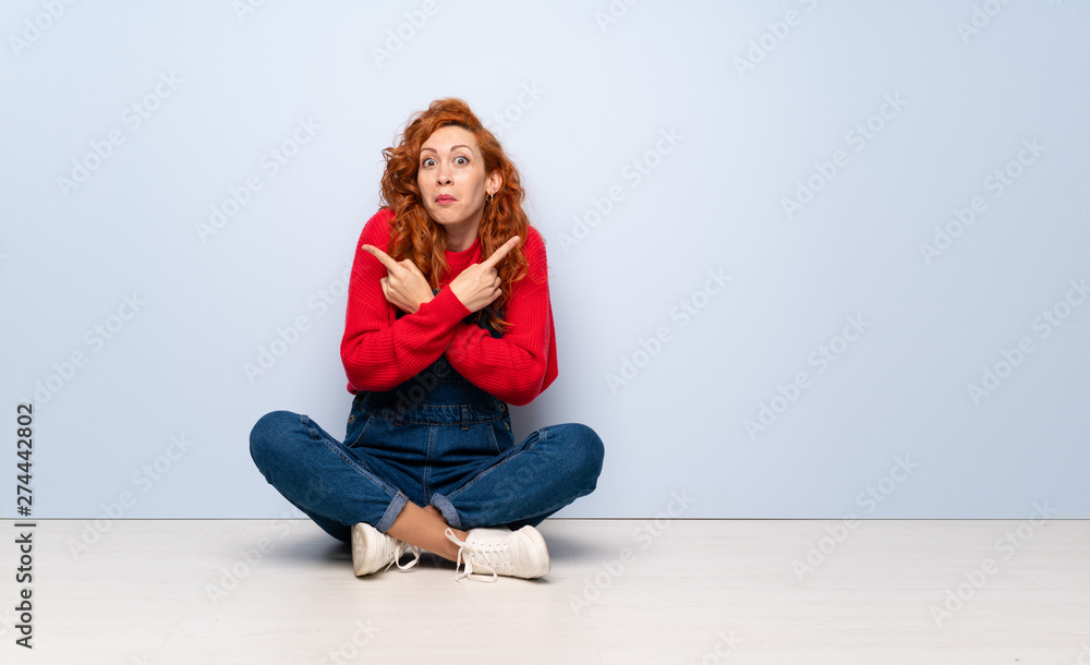 Redhead woman with overalls sitting on the floor pointing to the laterals having doubts Redhead woman with overalls sitting on the floor pointing to the laterals having doubts