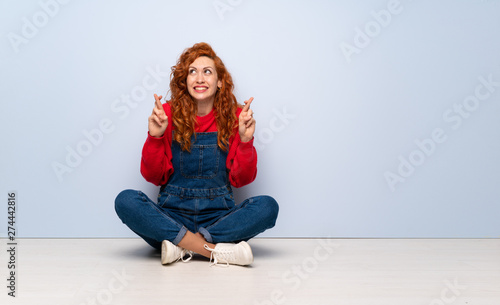Redhead woman with overalls sitting on the floor with fingers crossing and wishing the best