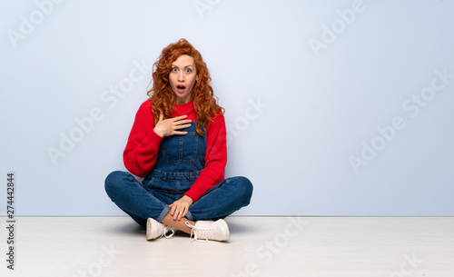 Redhead woman with overalls sitting on the floor surprised and shocked while looking right