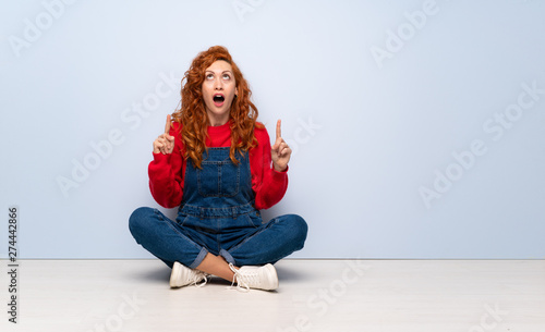 Redhead woman with overalls sitting on the floor surprised and pointing up