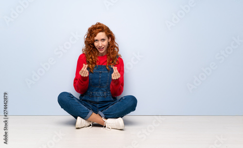 Redhead woman with overalls sitting on the floor making money gesture