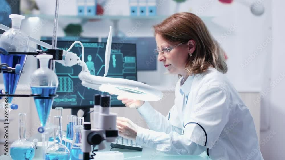 Chemist woman analysis a sample using a big magnifying glass. In the background - modern research facility with an active team