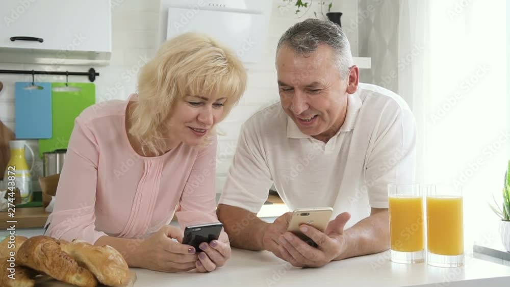 Senior family couple enjoying digital technology during breakfast in kitchen. Happy mature wife and elderly man holding smartphone and cellphone in hands and sharing video and photo in mobile app near Senior family couple enjoying digital technology during breakfast in kitchen. Happy mature wife and elderly man holding smartphone and cellphone in hands and sharing video and photo in mobile app near