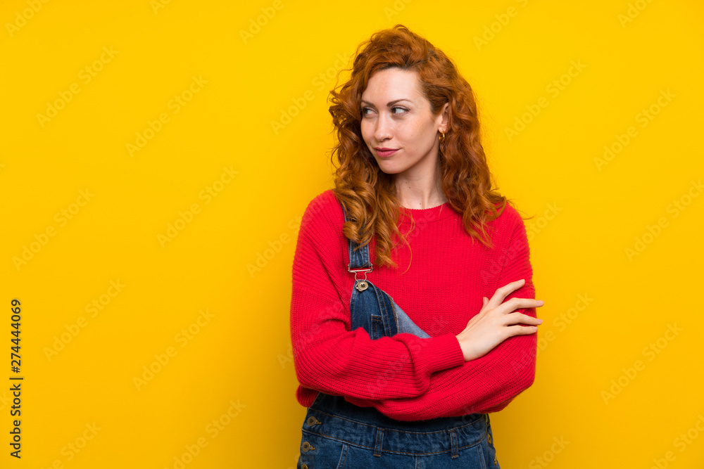 Redhead woman with overalls over isolated yellow wall standing and thinking an idea Redhead woman with overalls over isolated yellow wall standing and thinking an idea