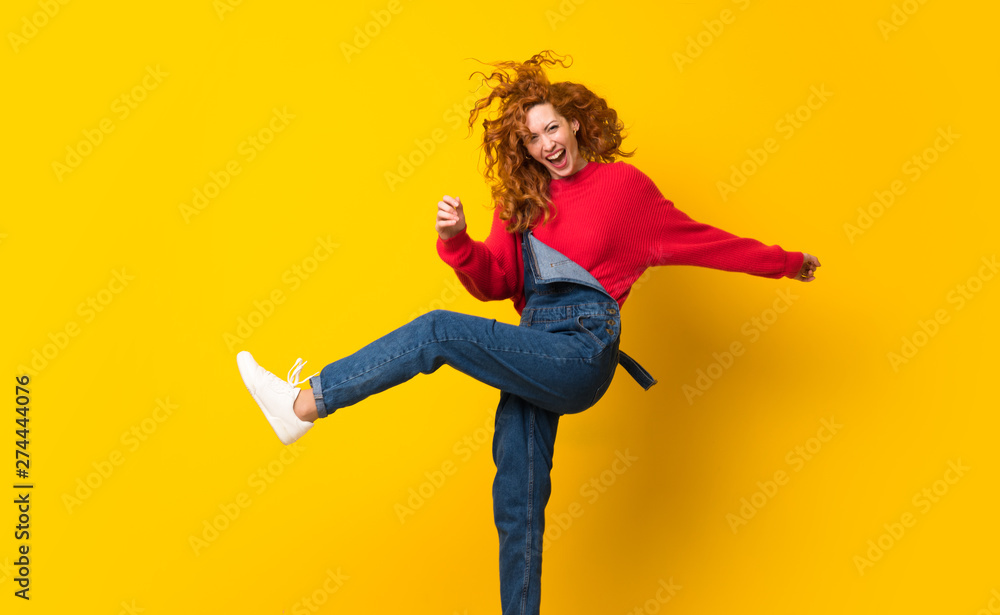 Redhead woman with overalls jumping over isolated yellow wall Redhead woman with overalls jumping over isolated yellow wall