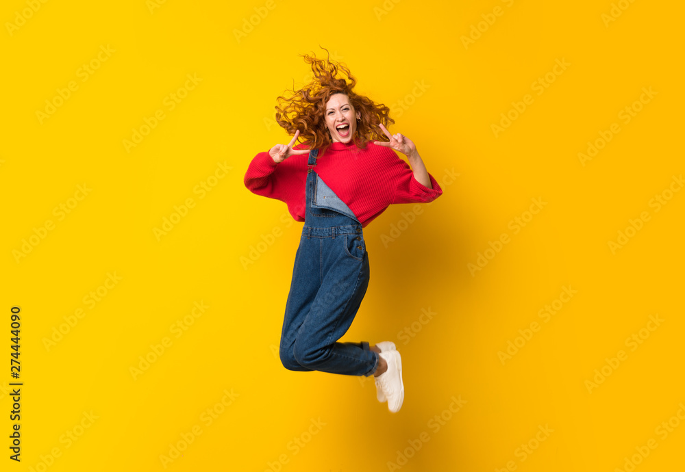 Redhead woman with overalls jumping over isolated yellow wall Redhead woman with overalls jumping over isolated yellow wall