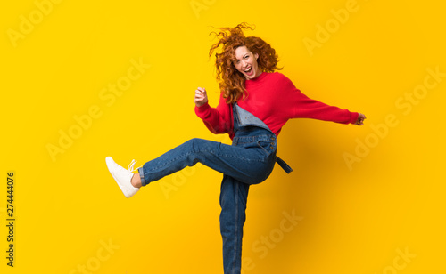 Redhead woman with overalls jumping over isolated yellow wall