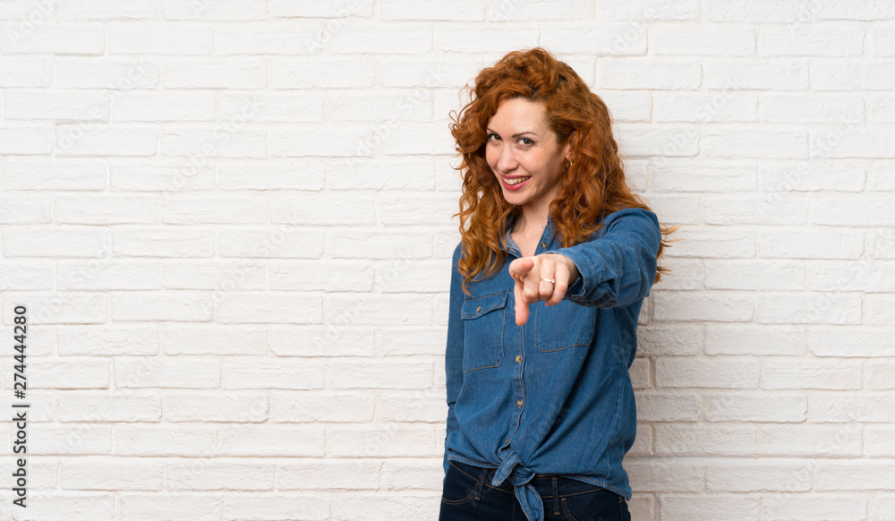Redhead woman over white brick wall points finger at you with a confident expression Redhead woman over white brick wall points finger at you with a confident expression