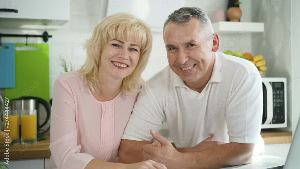 Happy family couple posing for photo in modern kitchen. Cheerful mature wife and senior husband standing at counter in cozy home interior and smiling. Happy family couple posing for photo in modern kitchen. Cheerful mature wife and senior husband standing at counter in cozy home interior and smiling.