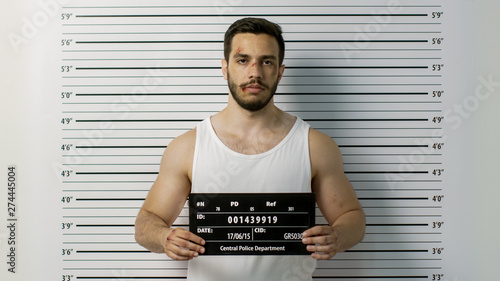 In a Police Station Arrested Beaten Man Poses for Front View Mugshot. He Wears Singlet, is Heavily Bruised and Holds Placard. Height Chart in the Background.