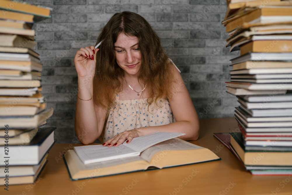 Tired female student reading among books. Pensive young woman sitting at table with pile of book and reading on background of grey wall Tired female student reading among books. Pensive young woman sitting at table with pile of book and reading on background of grey wall