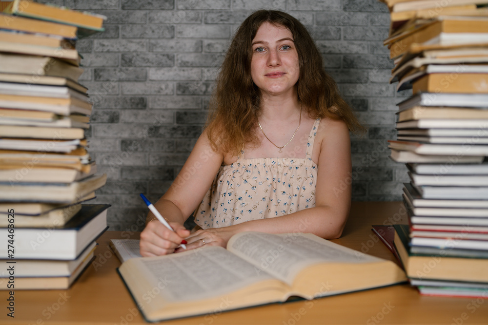 Tired female student reading among books. Pensive young woman sitting at table with pile of book and reading on background of grey wall Tired female student reading among books. Pensive young woman sitting at table with pile of book and reading on background of grey wall