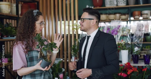 Cute florist friendly cheerful girl is describing beautiful green plants to bearded man in flowers shop standing together holding pots and talking. Business and people concept.