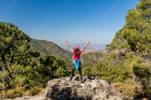 Senderista con los brazos extendidos en señal de victoria tras subir una montaña. Vista de una cadena montañosa al fondo. Cielo azul.
