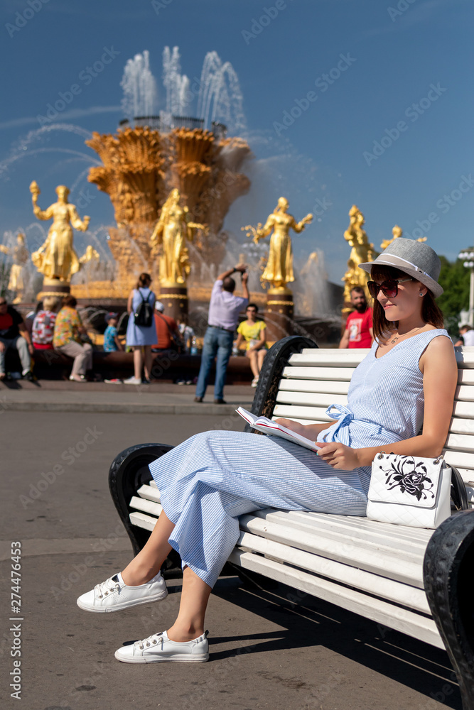 Obraz premium Happy young and stylish woman with hat and book sitting on the bench in the park.