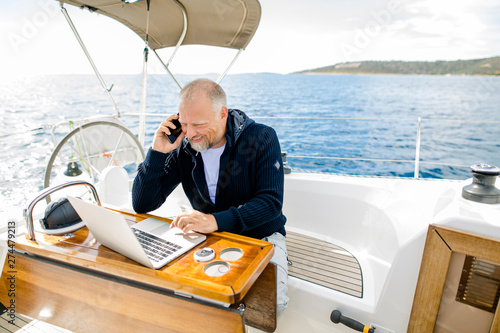 Digitaler Nomade arbeitet am Laptop lachend und telefonierend auf einem Segelboot