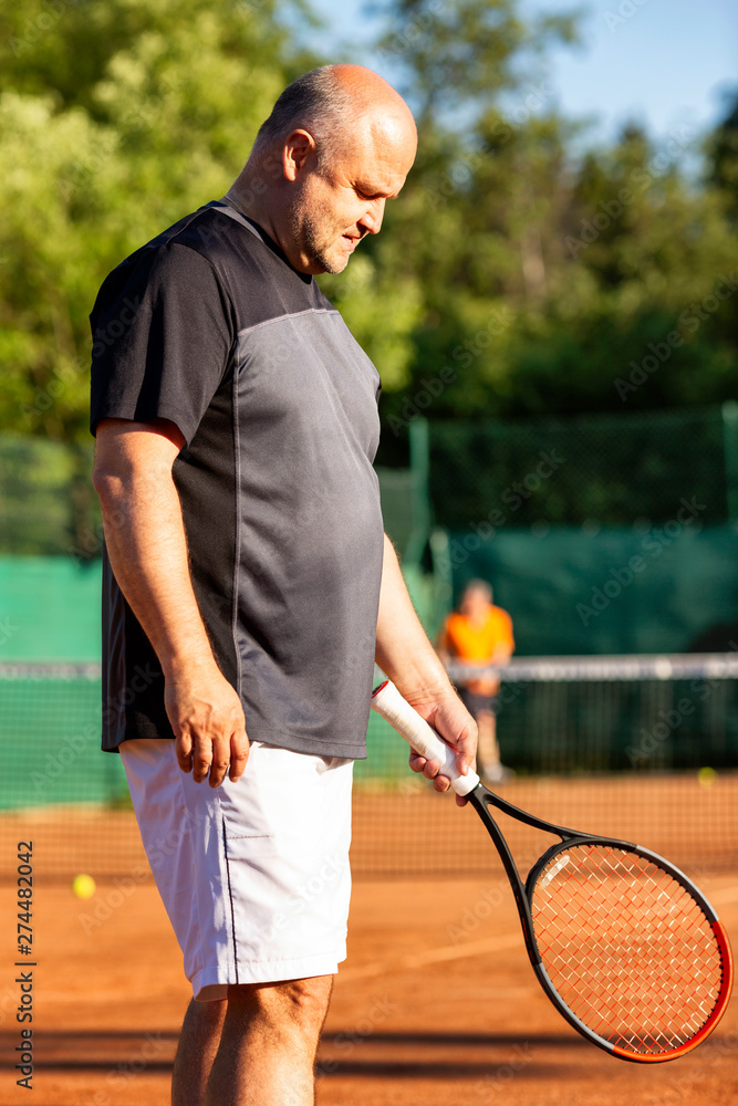 A middle-aged bald man plays tennis on the outdoor court. Sunny day. Vertical.