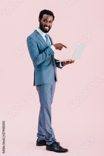 smiling african american businessman pointing at laptop isolated on pink