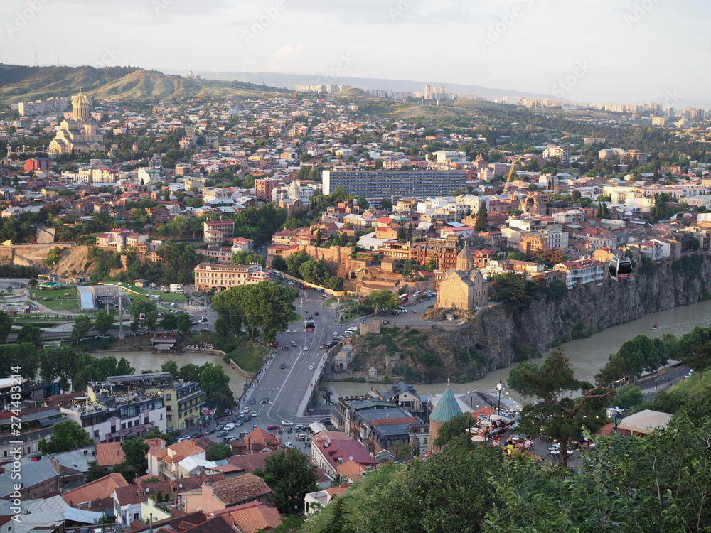 Aerial view from the Narikala fortress on the Tbilisi city, the capital of Georgia, full of small tiled roof houses, green trees, bridges over the Kura river