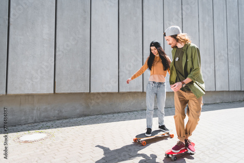 happy man holding hands with beautiful woman, riding on skateboard near concrete wall