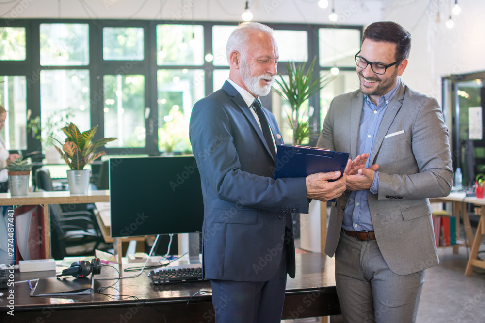 Φωτογραφία Senior businessman in formal wear discussing data from clipboard with younger male colleague while standing together in office